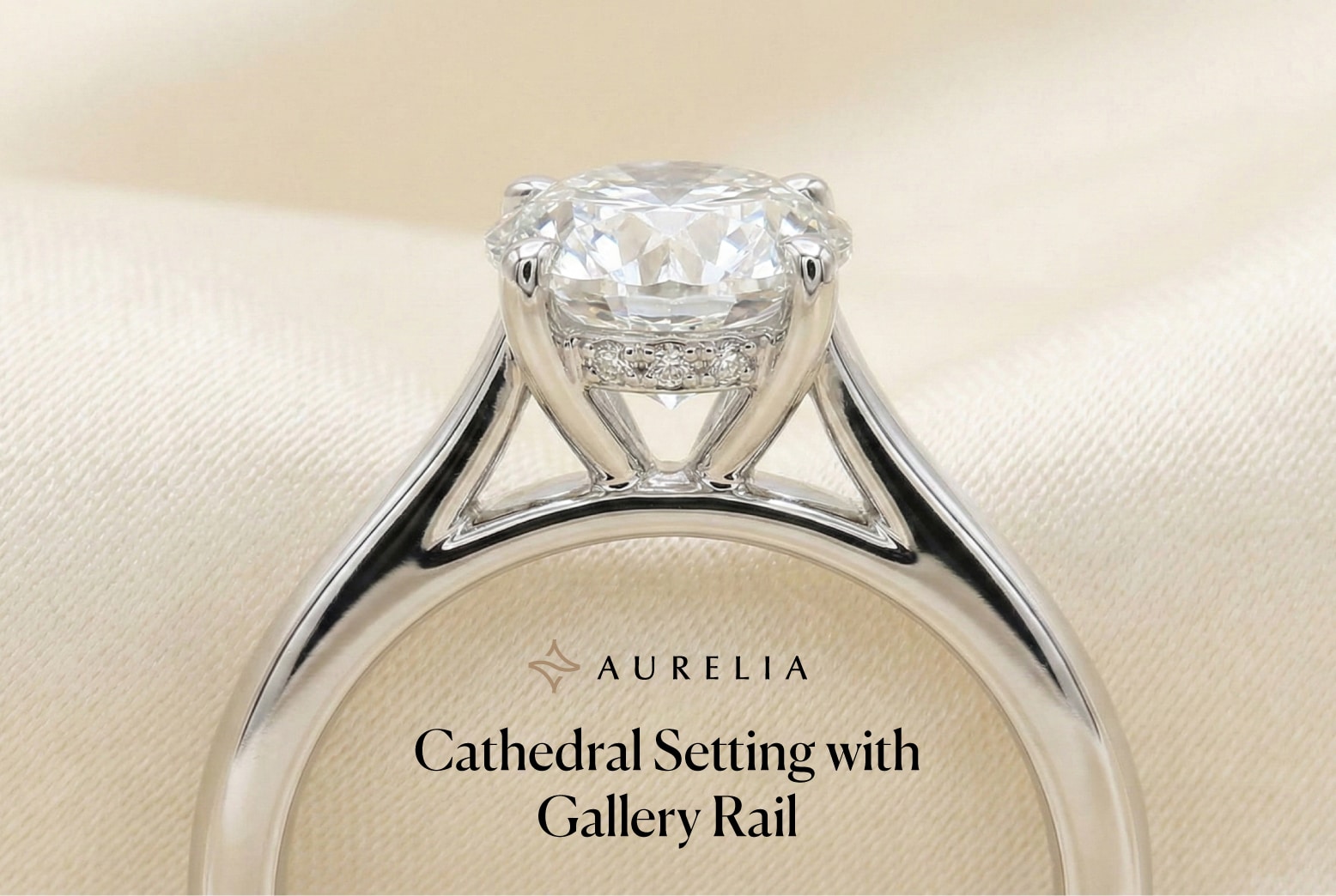 A close-up, profile view of an Aurelia engagement ring featuring a round brilliant-cut diamond in a cathedral setting. The ring is crafted in polished white gold or platinum, showcasing elegant arches that sweep up from the band to secure the center stone. A decorative hidden halo gallery rail, encrusted with small pavé-set diamonds, encircles the base of the prongs. The ring is set against a soft, cream-colored draped fabric background.