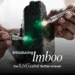 A close-up, dramatic image of a massive, rough emerald crystal named 'Imboo'. Two hands are shown holding what appear to be gemological tools or lights, illuminating the vibrant green facets of the raw emerald. The text on the image introduces the stone as "Imboo" and describes it as "the 11,685 carat 'Buffalo' emerald."