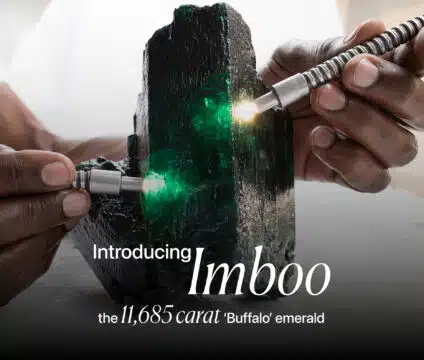 A close-up, dramatic image of a massive, rough emerald crystal named 'Imboo'. Two hands are shown holding what appear to be gemological tools or lights, illuminating the vibrant green facets of the raw emerald. The text on the image introduces the stone as "Imboo" and describes it as "the 11,685 carat 'Buffalo' emerald."
