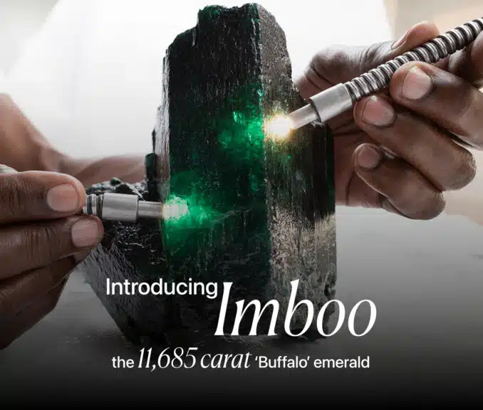 A close-up, dramatic image of a massive, rough emerald crystal named 'Imboo'. Two hands are shown holding what appear to be gemological tools or lights, illuminating the vibrant green facets of the raw emerald. The text on the image introduces the stone as "Imboo" and describes it as "the 11,685 carat 'Buffalo' emerald."