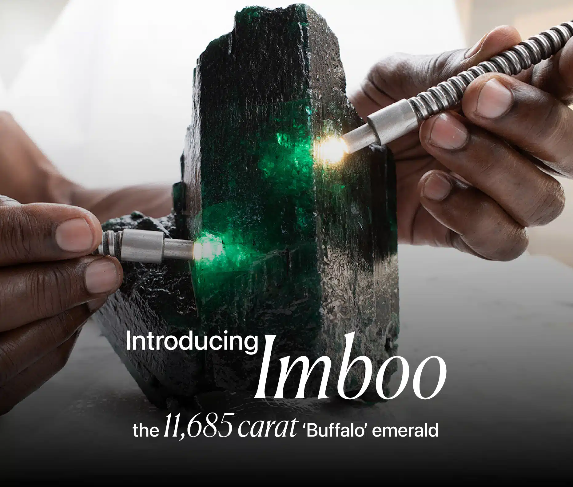 A close-up, dramatic image of a massive, rough emerald crystal named 'Imboo'. Two hands are shown holding what appear to be gemological tools or lights, illuminating the vibrant green facets of the raw emerald. The text on the image introduces the stone as "Imboo" and describes it as "the 11,685 carat 'Buffalo' emerald."