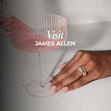 A close-up shot focuses on a woman's hand, adorned with an elegant gold ring featuring a large marquise-cut diamond and a matching gold diamond band. Her hand rests on a white surface where pink liquid is spilling from a ribbed pink coupe glass tilted in the upper left of the frame. White text overlays the image, reading "Visit JAMES ALLEN™".
