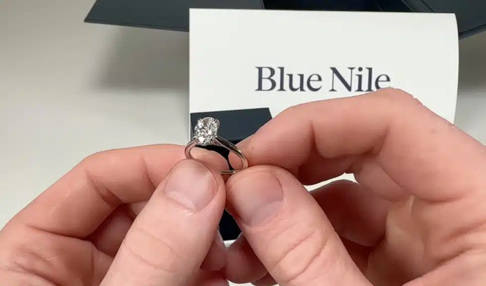A close-up of the Blue Nile oval diamond engagement ring held by hand, with the official brand card visible in the background.