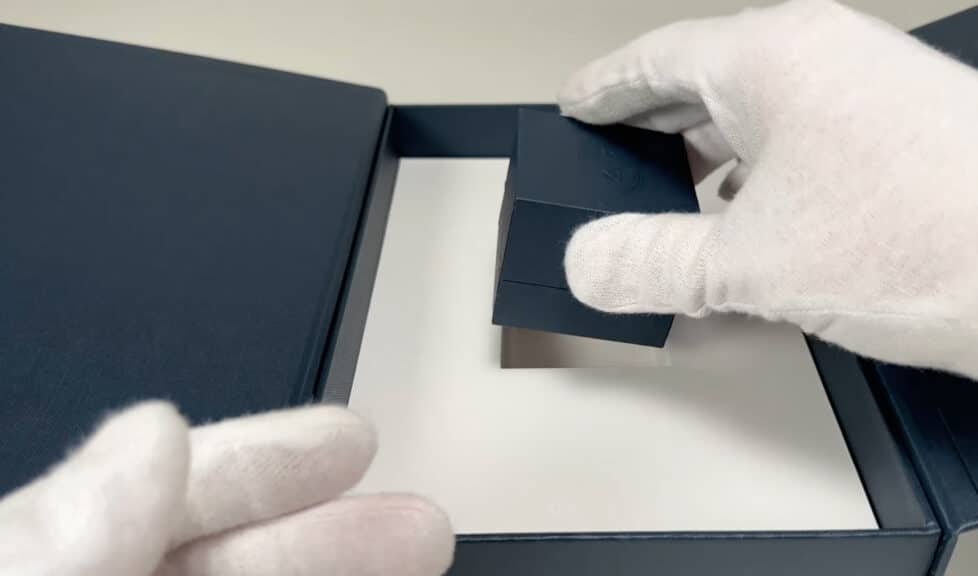 A gloved hand carefully lifts the small, dark blue ring box out of its custom cutout within the larger Blue Nile package.