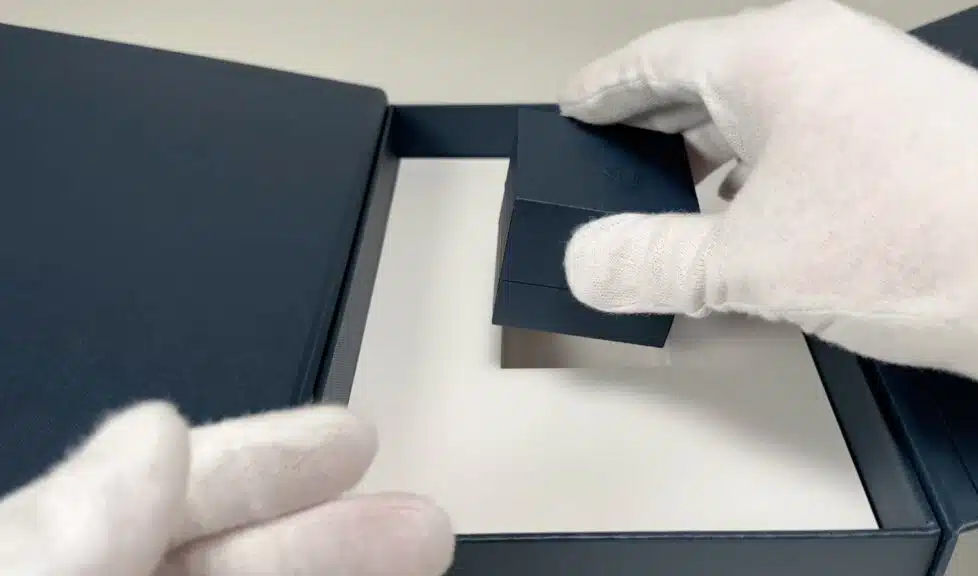 A gloved hand carefully lifts the small, dark blue ring box out of its custom cutout within the larger Blue Nile package.