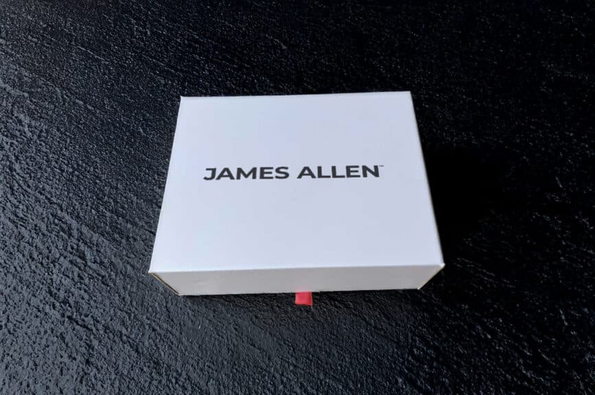 A pristine white James Allen jewelry box featuring the new James Allen logo, resting on a dark, textured surface, representing the arrival and packaging of a James Allen order.