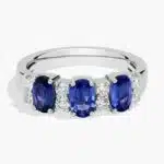 A 14K White Gold 3 Stone Diamond Rings With Sapphires ring (3.60 mm wide) featuring three Oval Blue Sapphires set in prongs, accented by 0.31 CTW Round Diamonds on the shank.