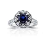 A 14K White Gold Antique Style Vintage Art Deco Sapphire Rings engagement ring featuring a 1.3 Carat Round Blue Sapphire center stone, a Floral Design, and a 1/5 CTW Pave set diamond halo.