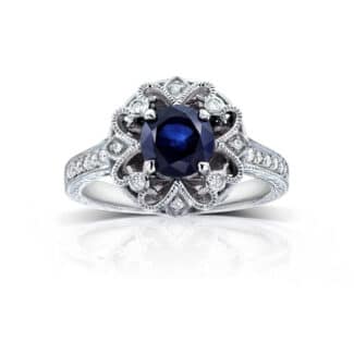 A 14K White Gold Antique Style Vintage Art Deco Sapphire Rings engagement ring featuring a 1.3 Carat Round Blue Sapphire center stone, a Floral Design, and a 1/5 CTW Pave set diamond halo.