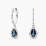 A pair of 14K White Gold Sapphire and Diamond Dangle Earrings featuring two 6x4 mm Pear Shaped Blue Sapphires (1 CT total) and a 0.22 CTW Round Diamond Halo, secured with Hinged Snap Backs.