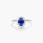 An 18K White Gold Oval Halo Engagement Rings With Diamonds and Sapphires ring featuring a 1.05 Carat Oval Blue Sapphire center stone and a 0.27 Carat Total Weight round diamond halo.