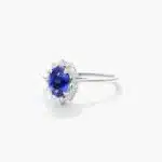 An 18K White Gold Oval Halo Engagement Rings With Diamonds and Sapphires ring featuring a 1.05 Carat Oval Blue Sapphire center stone and a 0.27 Carat Total Weight round diamond halo.