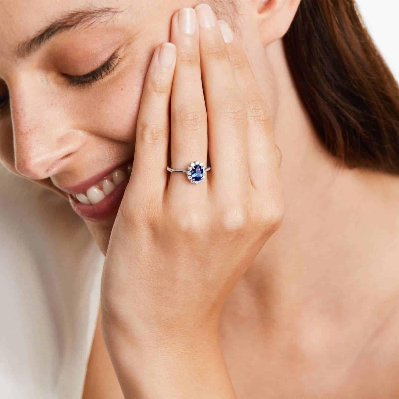 An 18K White Gold Oval Halo Engagement Rings With Diamonds and Sapphires ring featuring a 1.05 Carat Oval Blue Sapphire center stone and a 0.27 Carat Total Weight round diamond halo.