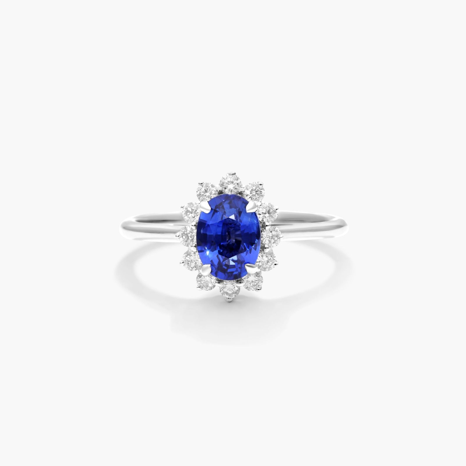 An 18K White Gold Oval Halo Engagement Rings With Diamonds and Sapphires ring featuring a 1.05 Carat Oval Blue Sapphire center stone and a 0.27 Carat Total Weight round diamond halo.