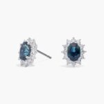 An 18K White Gold White Gold and Sapphire Earrings set featuring two 7x5 mm Oval Blue Sapphires surrounded by a 0.72 CTW Round Diamond Halo, secured with Push Backs.