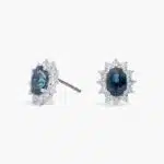 An 18K White Gold White Gold and Sapphire Earrings set featuring two 7x5 mm Oval Blue Sapphires surrounded by a 0.72 CTW Round Diamond Halo, secured with Push Backs.