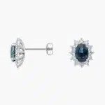 An 18K White Gold White Gold and Sapphire Earrings set featuring two 7x5 mm Oval Blue Sapphires surrounded by a 0.72 CTW Round Diamond Halo, secured with Push Backs.