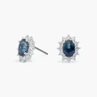 An 18K White Gold White Gold and Sapphire Earrings set featuring two 7x5 mm Oval Blue Sapphires surrounded by a 0.72 CTW Round Diamond Halo, secured with Push Backs.