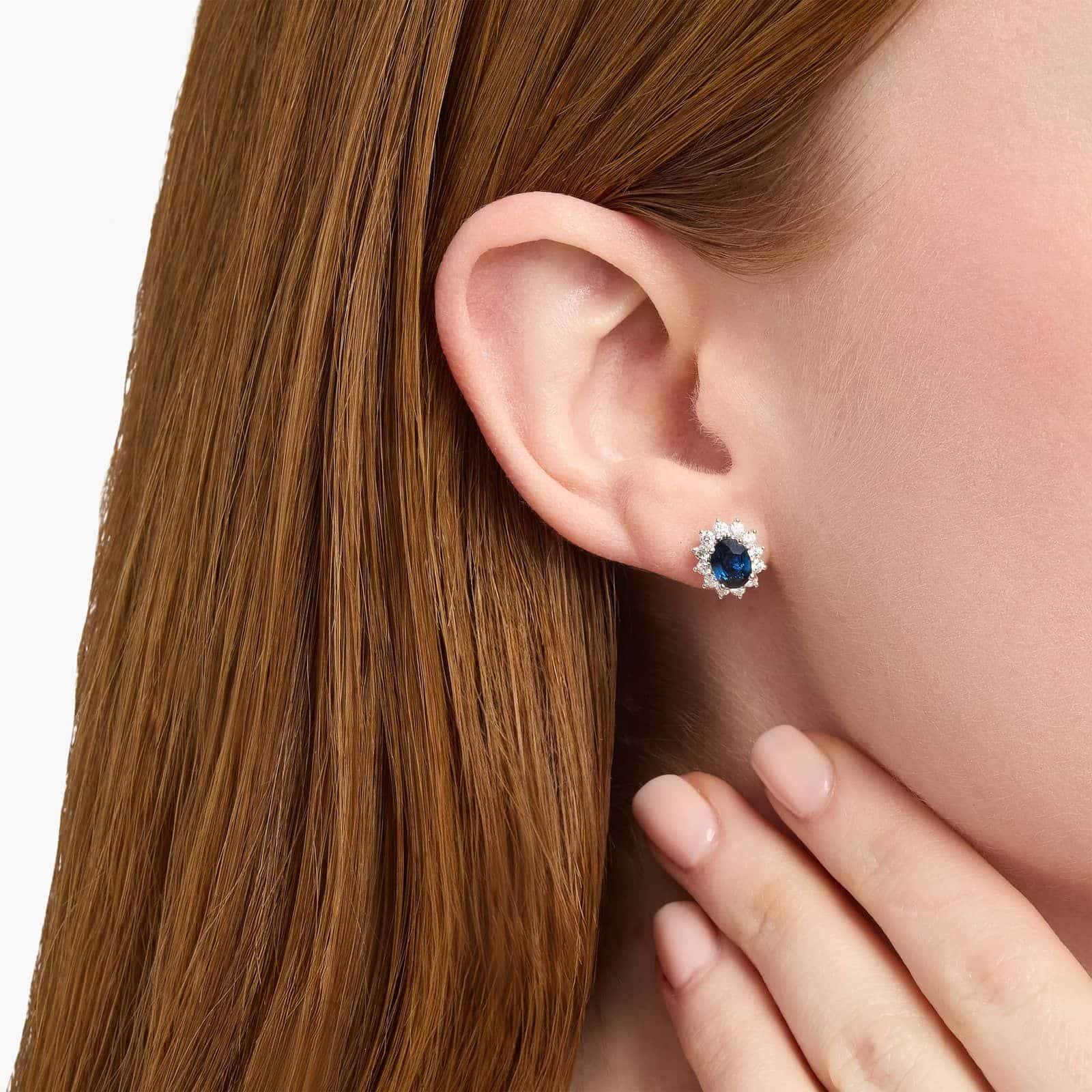 oval-sapphire-diamond-halo-white-gold-and-sapphire-earrings-18k-0-72ctw model view An 18K White Gold White Gold and Sapphire Earrings set featuring two 7x5 mm Oval Blue Sapphires surrounded by a 0.72 CTW Round Diamond Halo, secured with Push Backs.